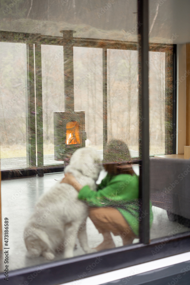 Young woman with her dog at modern house on nature. Girl in green sweater sitting near the window an