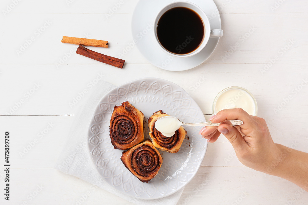 Woman adding cream onto tasty cinnamon rolls on white wooden background