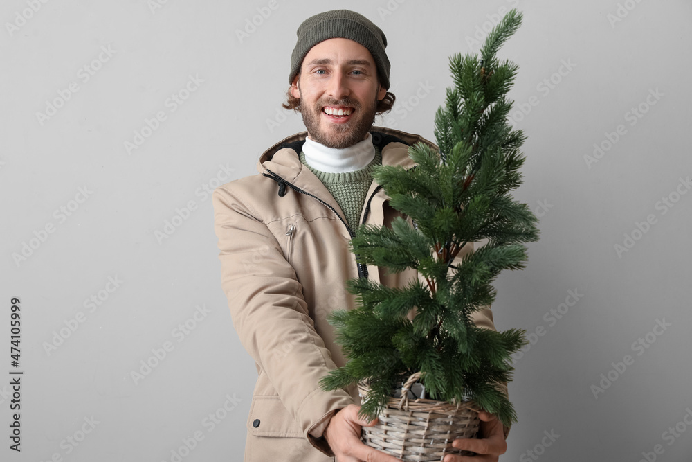 Stylish young man in winter clothes and with Christmas tree on light background