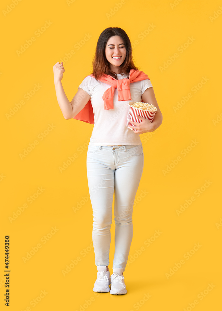 Happy young Asian woman with bucket of tasty popcorn on yellow background