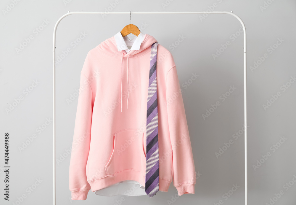 Rack with stylish pink hoodie, shirt and necktie near light wall