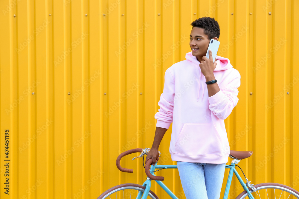 African-American teenage boy with bicycle talking by mobile phone near yellow fence