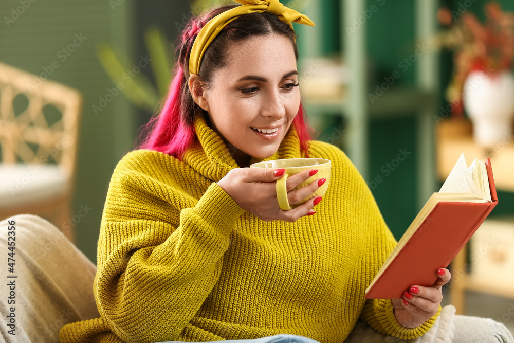 Beautiful woman in warm sweater with cup of tea reading book on sofa at home