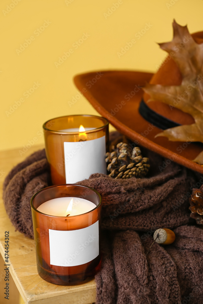 Burning candles in holders, scarf and autumn decor on table near yellow wall