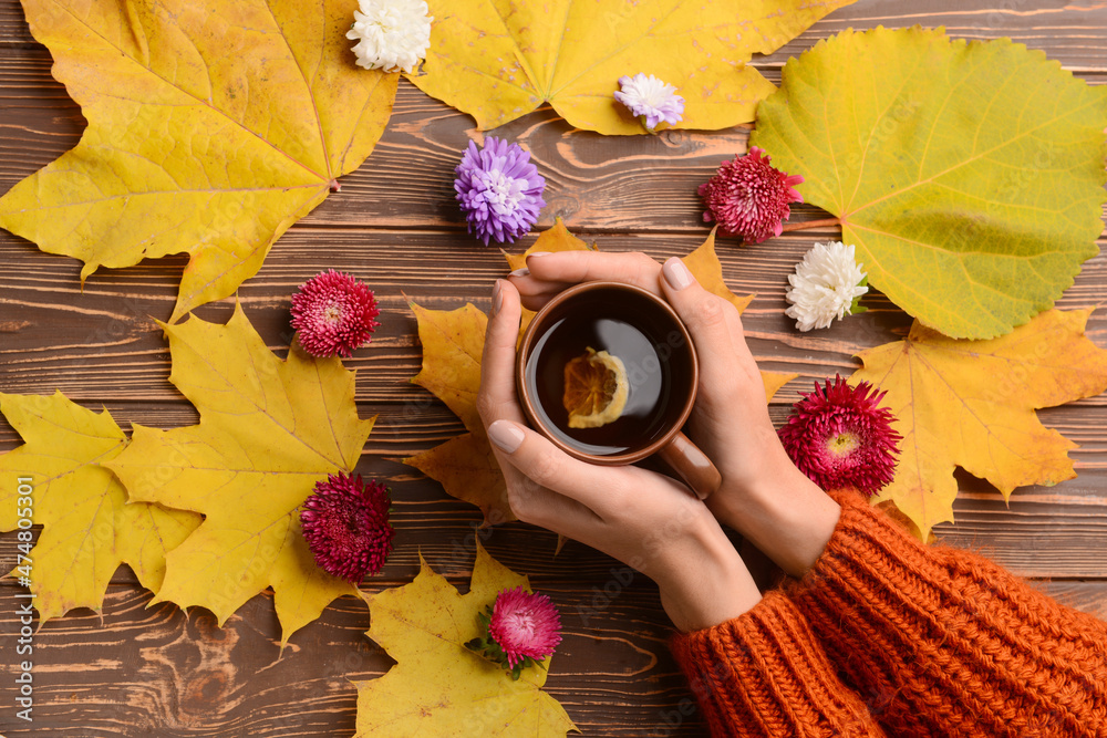 Female hands with cup of tea, autumn leaves and aster flowers on wooden background, closeup