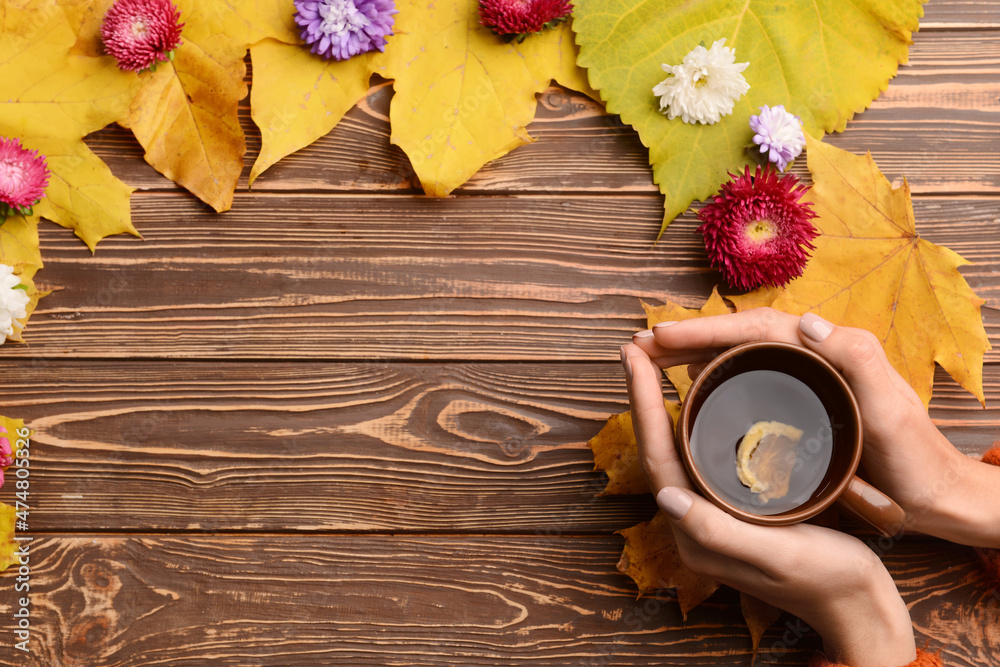 Female hands with cup of tea, autumn leaves and aster flowers on wooden background, closeup