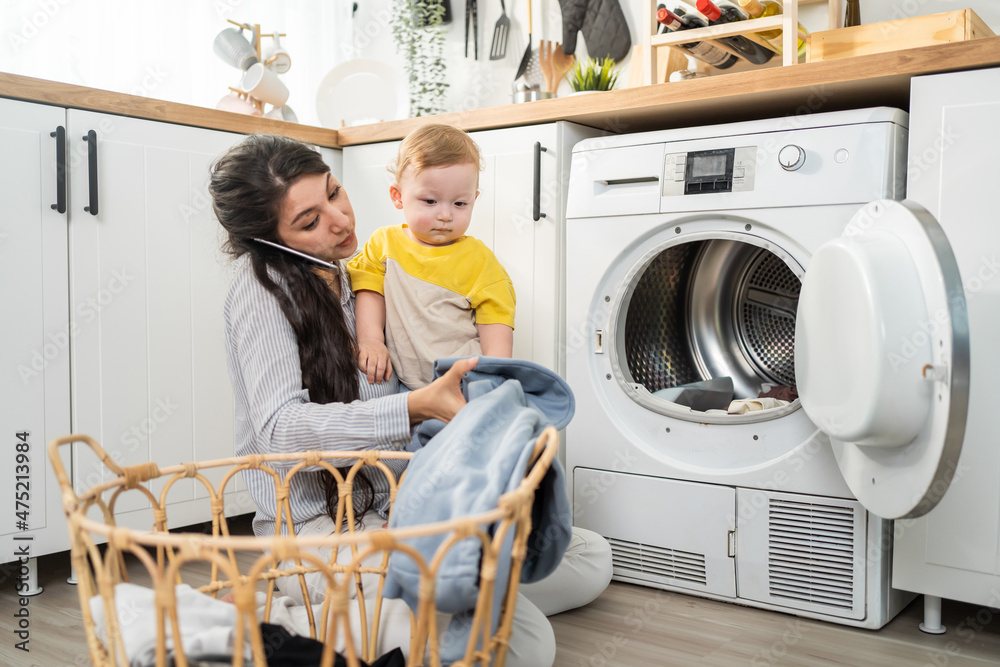 Caucasian busy mother doing housework with baby boy toddler in kitchen