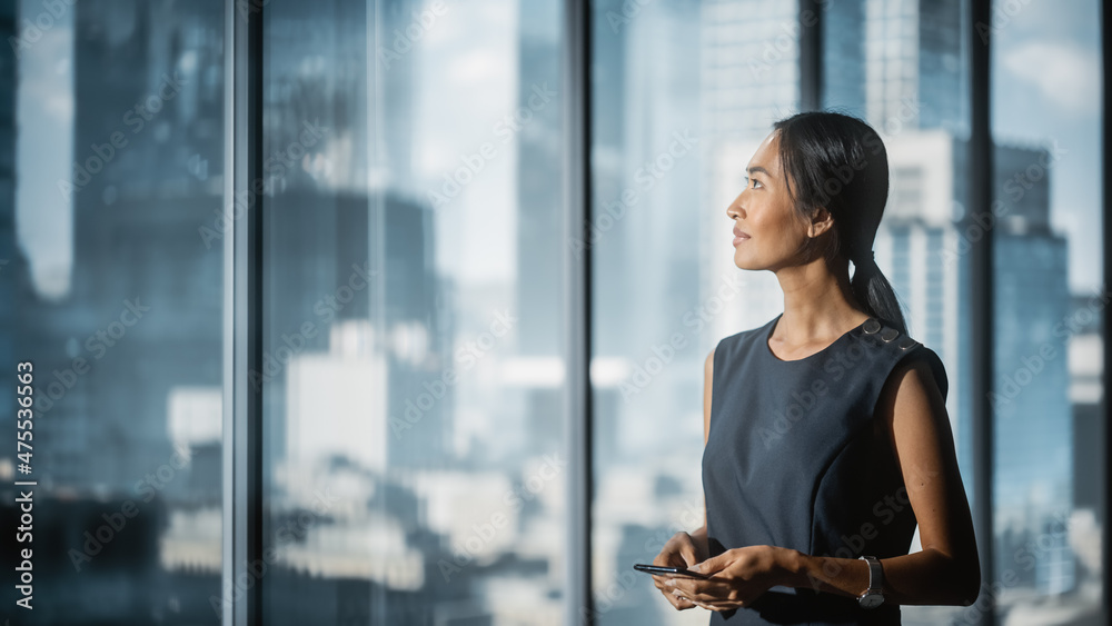 穿着时髦连衣裙的成功女商人站在现代办公室，使用智能手机，向外看