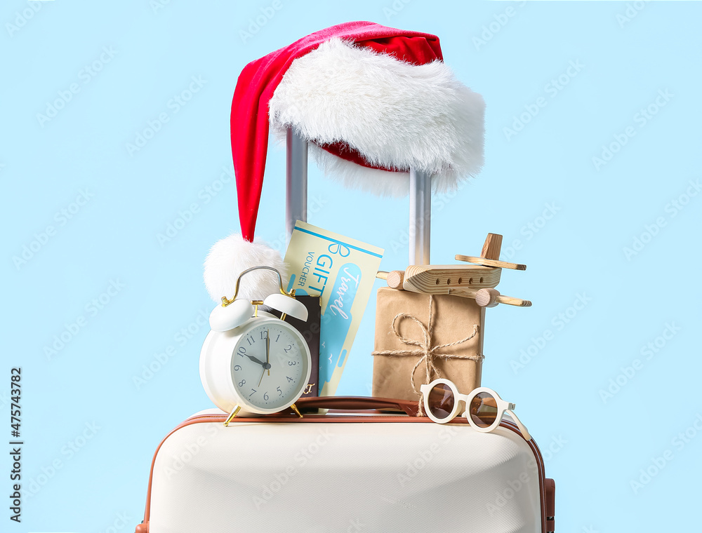 Suitcase with Christmas gift box, wooden airplane, alarm clock, sunglasses and Santa hat on blue bac