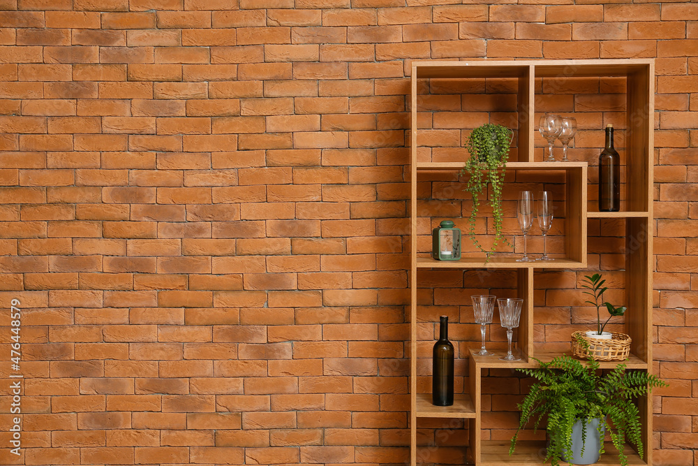 Shelving unit with bottles of wine, glasses and houseplants near brick wall