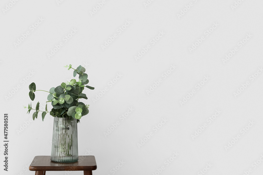 Vase with eucalyptus on wooden stool near light wall