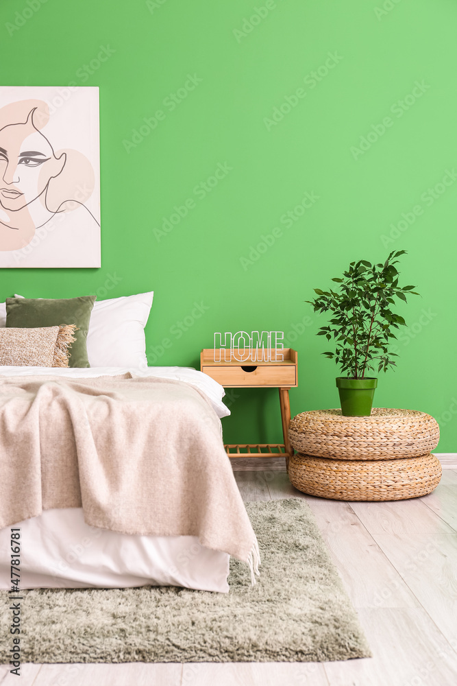 Comfortable bed, wooden table, houseplant and rattan poufs near green wall