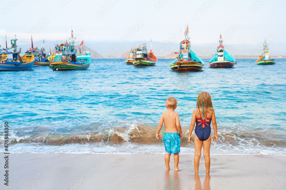 Happy family - kids walk along sea surf on sand beach, look at traditional fisher boats. Pantai Tanj