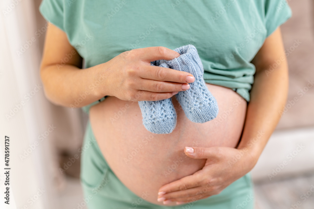 Expecting woman standing at the window and holding baby socks on her tummy. Lady holding her one han