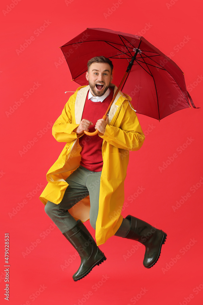 Jumping young man in stylish raincoat and with umbrella on color background