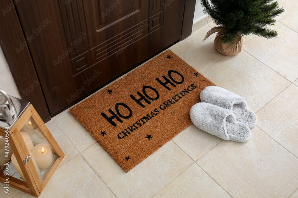 Mat with Christmas greeting, slippers, candles and small fir tree near dark wooden door