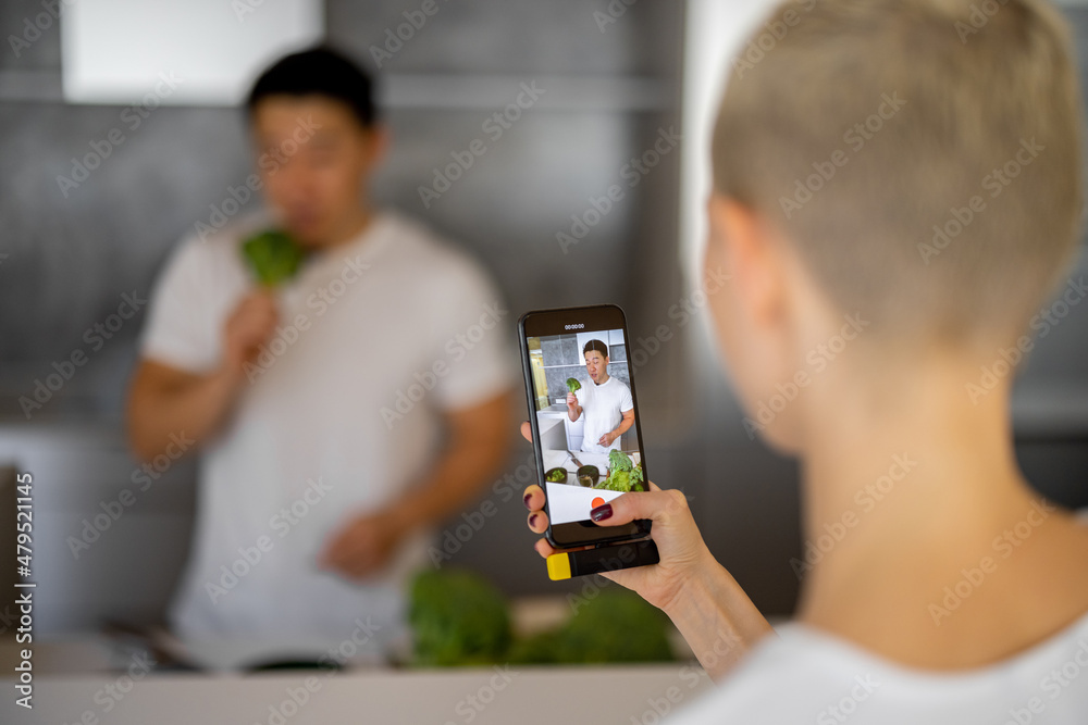 一名女子在智能手机上拍摄亚洲男子在厨房烹饪素食沙拉的视频。视频的概念