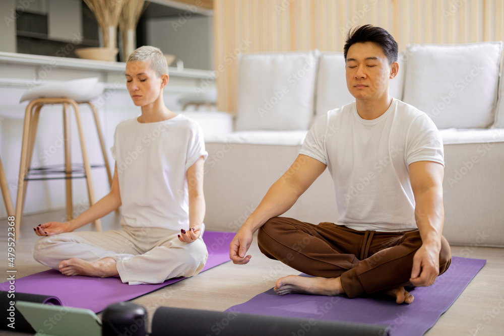 Multiracial couple practising yoga and meditating while sitting on fitness mats at home. Concept of 