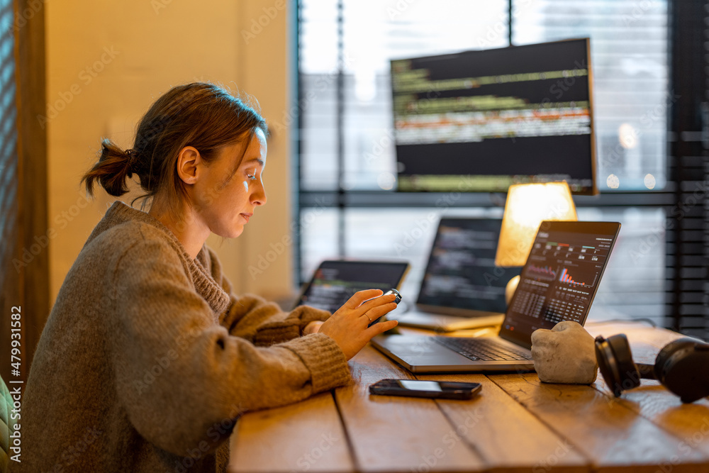 Young woman using phone while sitting at workplace with laptop and desktop computers at cozy home of