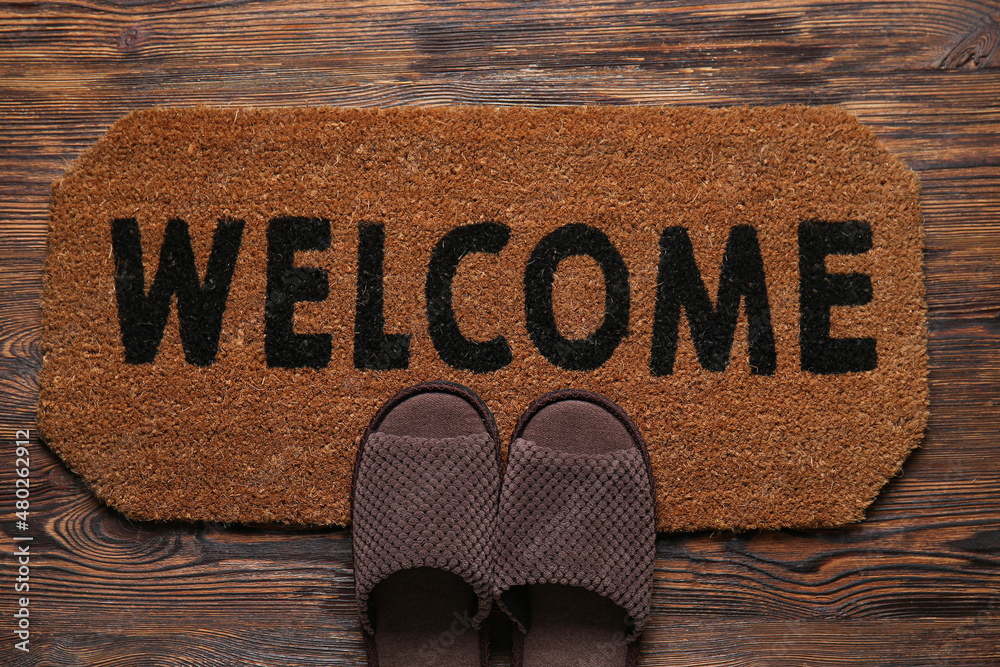 Door mat with word WELCOME and slippers on dark wooden floor