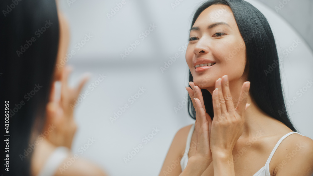 Portrait of Beautiful Asian Woman Gently Applying Face Cream Looking in Bathroom Mirror. Young Adult