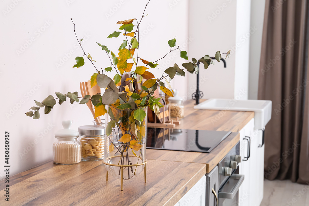 Vase with beautiful autumn branches and jars with food on wooden counter in light kitchen