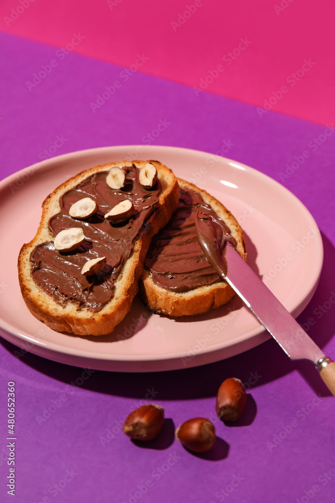 Plate of bread with chocolate paste and hazelnuts on color background