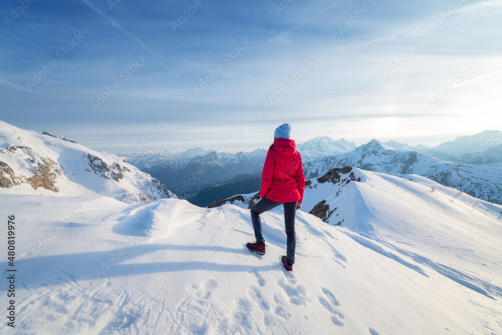 冬天日落时，雪山上穿着红色夹克的年轻女子。风景与美丽的女孩在一起