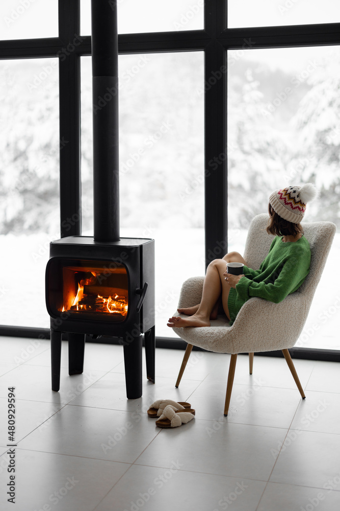 Woman sitting with cup on chair by the fireplace at modern house on nature during winter time. Conce