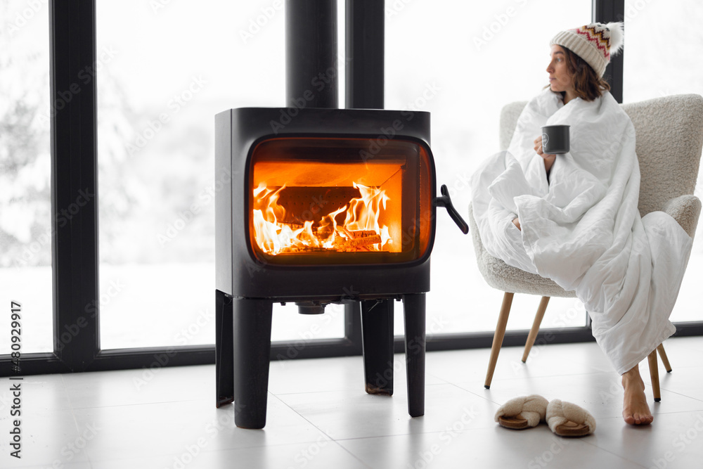 Woman covered with blanket enjoys winter time at home with burning fireplace, looking outside the wi