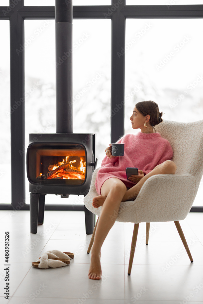 Woman sitting with cup on chair by the fireplace at modern house on nature during winter time. Conce