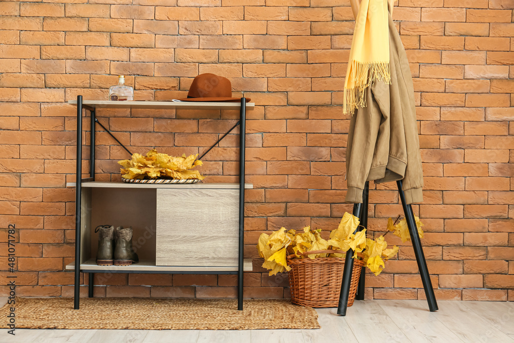 Hanger with jacket and basket with autumn leaves near brick wall in hallway