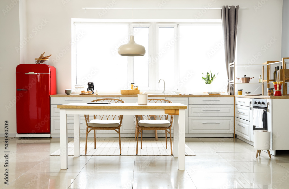 Interior of light modern kitchen with red fridge, white counters and dining table