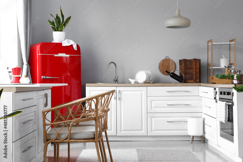 Interior of light modern kitchen with red fridge, white counters and wicker chairs