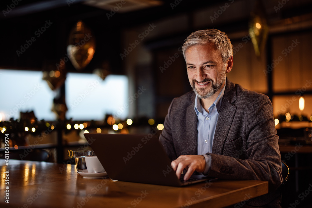 Smiling adult man, typing fast on his laptop keyboard, sitting in a bar.