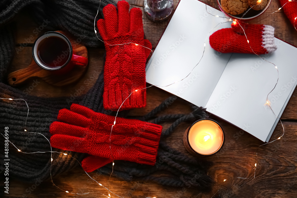 Warm gloves, scarf, cup of tea, blank book and burning candle on wooden background