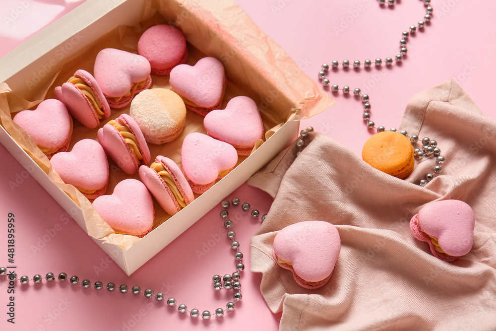 Box with tasty heart-shaped macaroons and beads on pink background