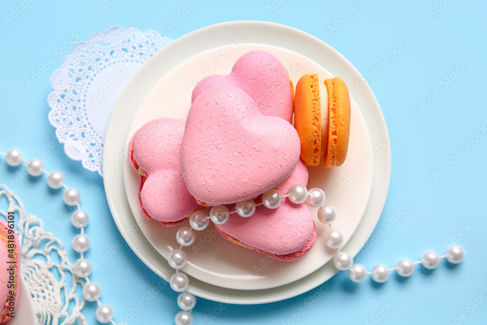 Plate with tasty heart-shaped macaroons and beads on blue background