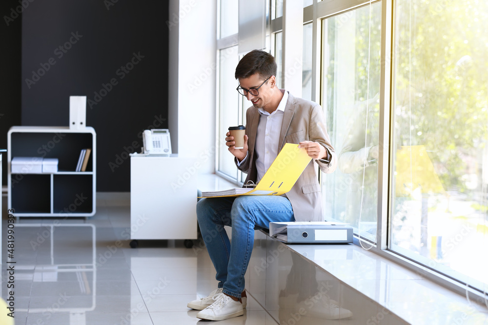 Young man with open folder and cup of coffee sitting on windowsill in office