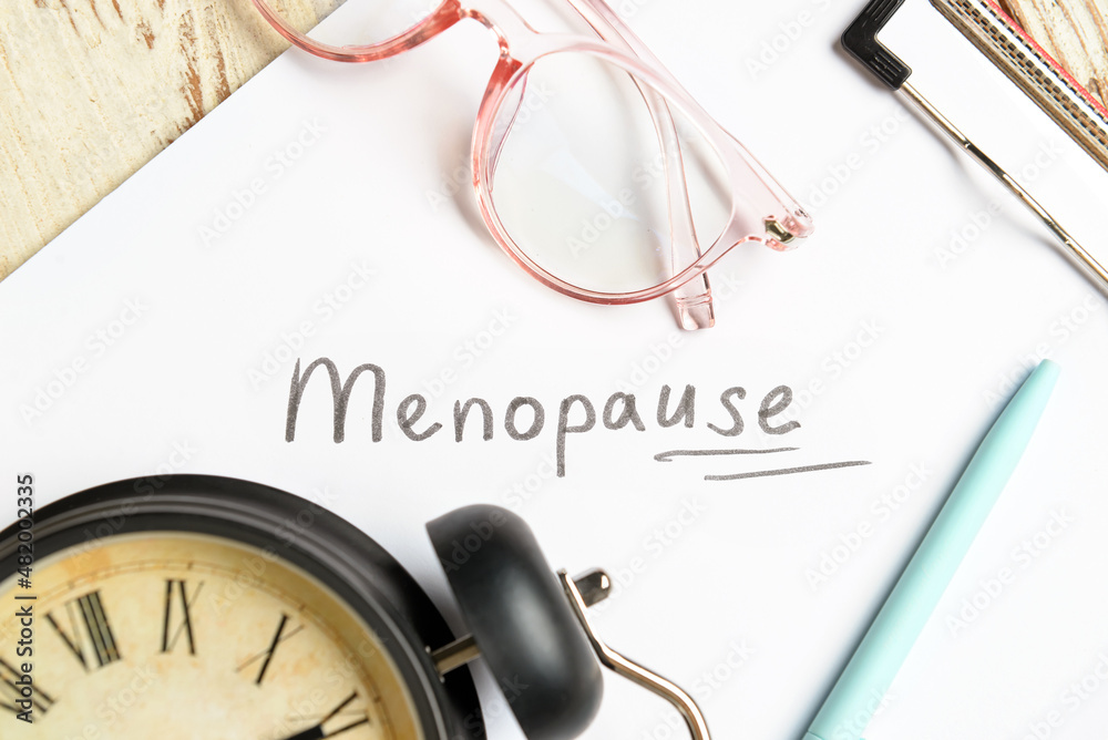 带有单词MENOPAUSE的剪贴板，浅色木背景上的眼镜和闹钟，特写