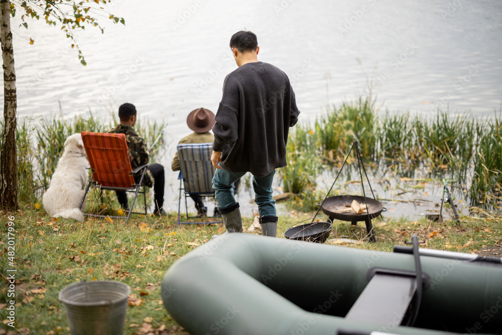 Asian man walks from rubber boat while his male multiracial friends fishing on river or lake coast. 