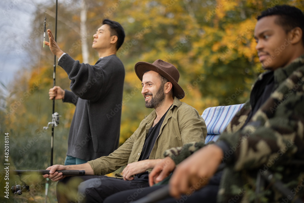 Multiracial male friends resting, talking and fishing on river or lake coast. Concept of leisure, ho