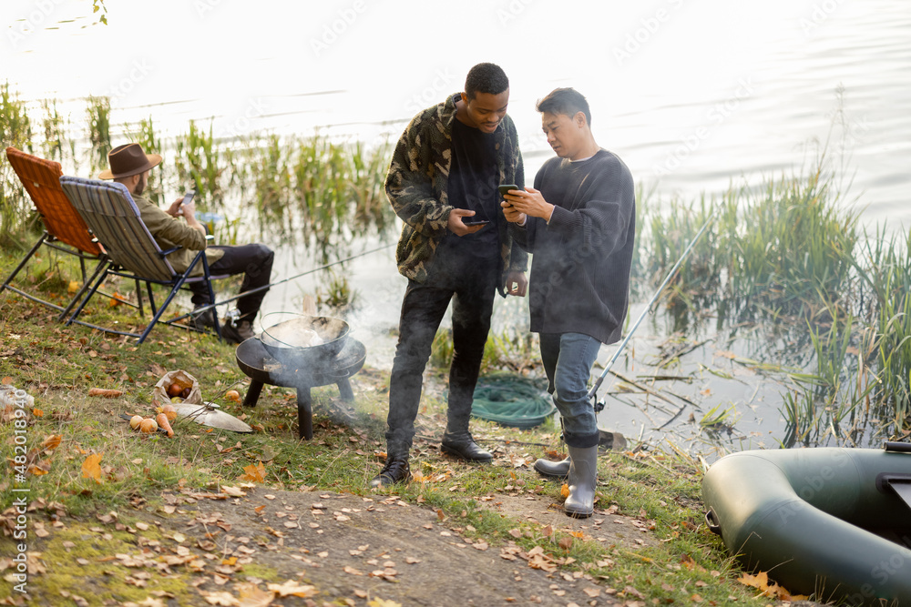 Asian and latin men using smartphones during resting on river or pond coast. Men fishing and cooking