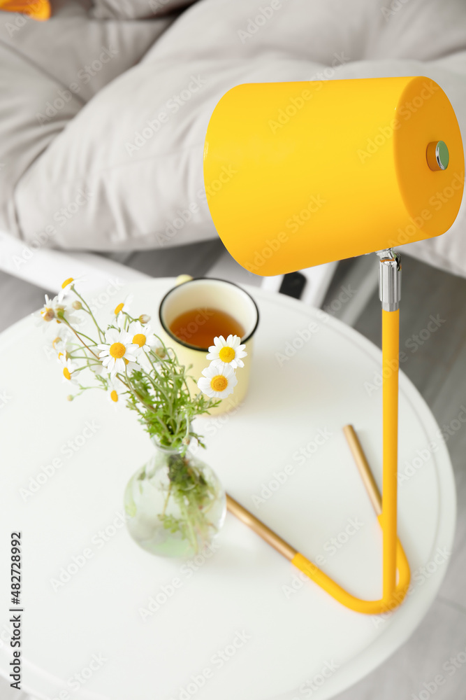 Modern lamp, vase with chamomile flowers and cup of tea on table