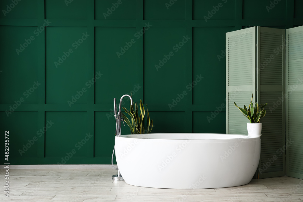 White bathtub with houseplants and folding screen near green wall