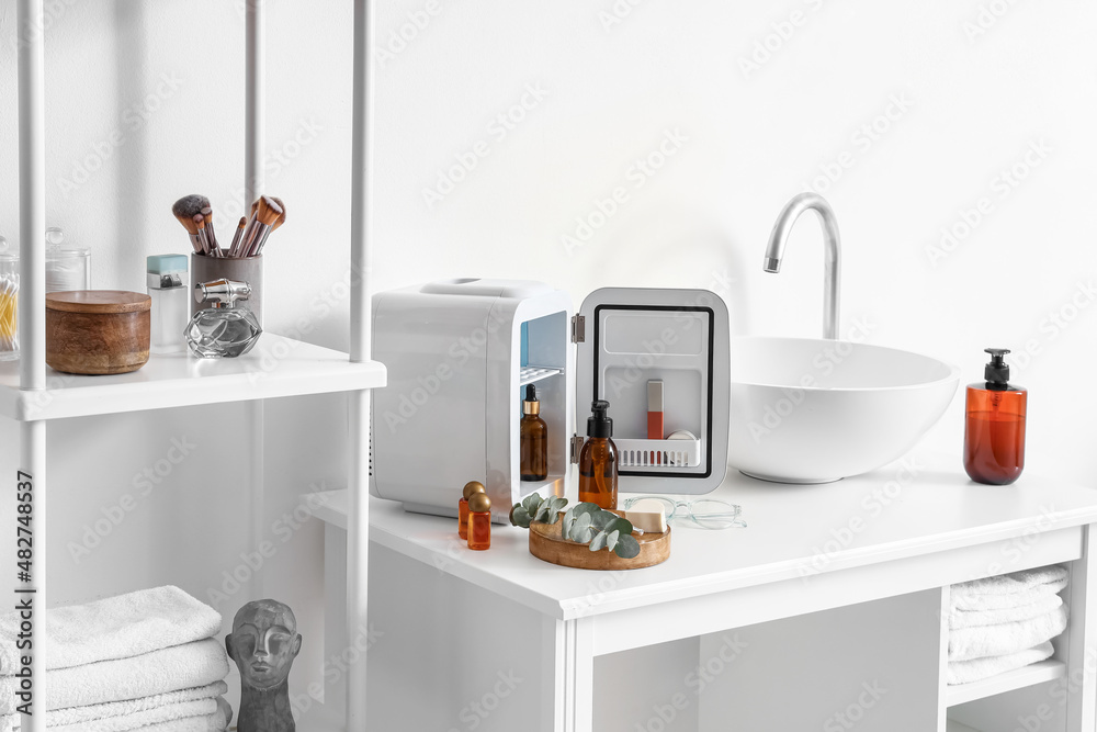 Sink, open refrigerator and cosmetic products on table near light wall in bathroom