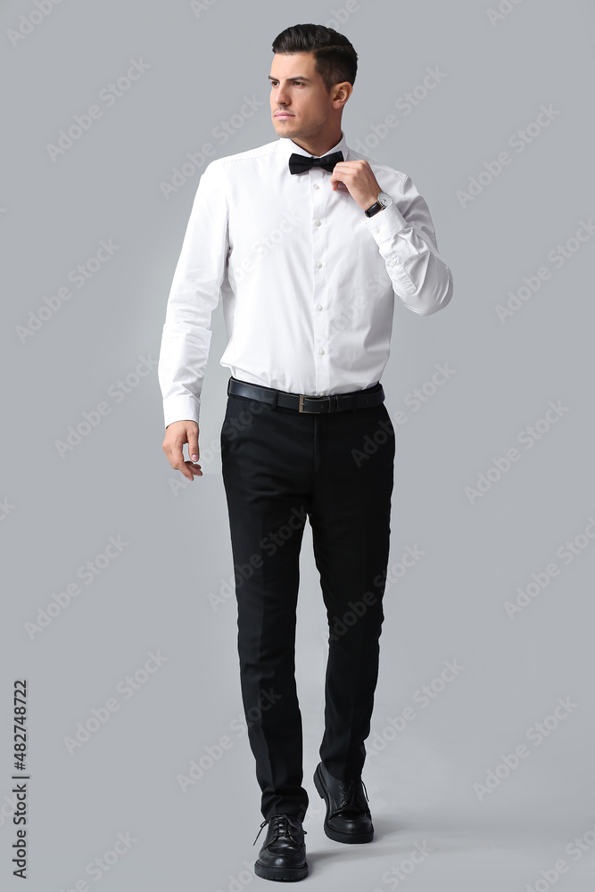 Fashionable gentleman looking aside and adjusting bow tie on grey background