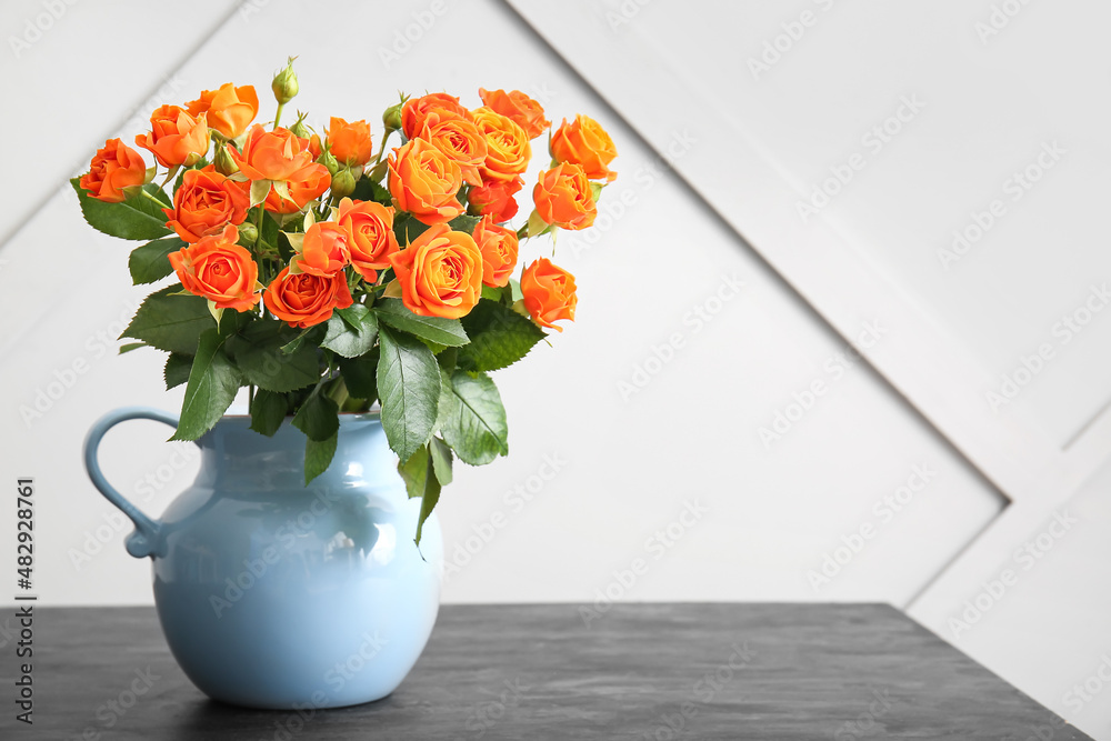 Jug with beautiful orange roses on table against light wall