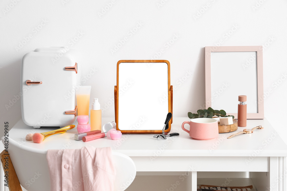 Table with cosmetic refrigerator, products and mirror near light wall