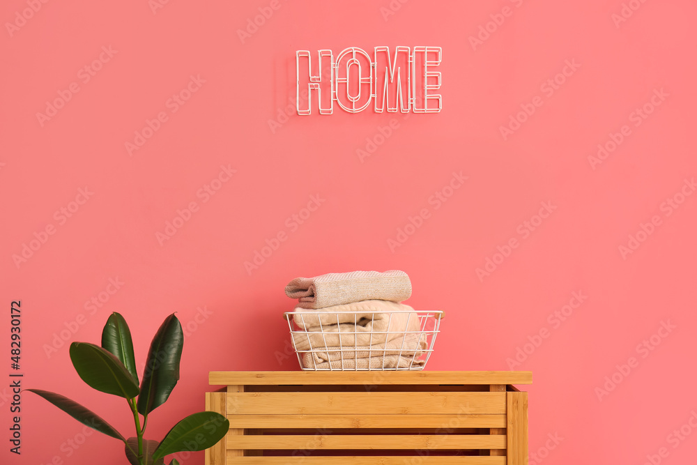 Basket with childrens sweaters on wooden table, houseplant and word HOME on color wall